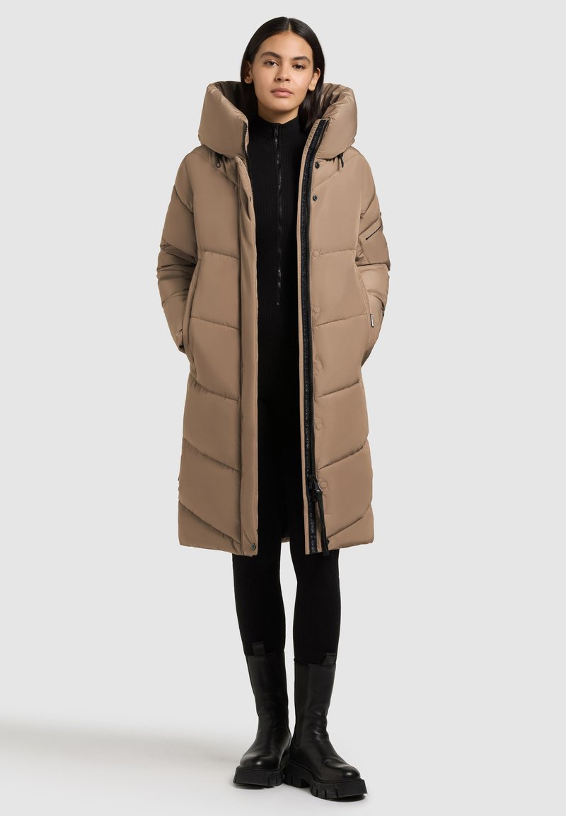 Kvinde iført en lang beige pufferfrakke med høj krave, sort ribbet outfit og sorte klodsede støvler, stående mod en lys baggrund.