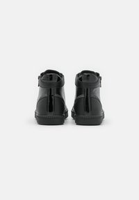 Paire de baskets noires montant à la cheville avec des panneaux texturés et brillants, fermeture éclair visible sur le côté extérieur, vues de dos.