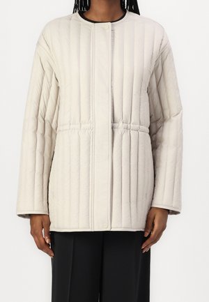Personne portant une veste matelassée beige clair avec des coutures verticales et un pantalon noir, visible des épaules jusqu'au milieu de la cuisse.