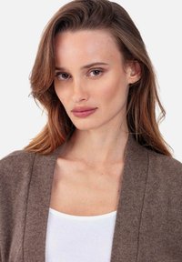 Cardigan en tricot marron à ouverture devant, avec une texture douce. Le modèle porte un débardeur blanc en dessous. Fond neutre.