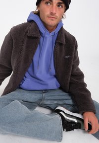 Giacca in pile marrone, felpa viola chiaro, jeans blu e scarpe sportive nere. Modello seduto, indossando un berretto nero aderente con un logo.