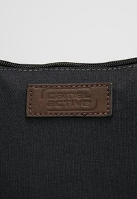 Gros plan sur un tissu noir avec une étiquette en cuir marron cousue embossée "camel active" sous une fermeture éclair noire.