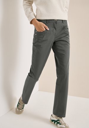 Personne portant un pantalon gris foncé à jambes droites, un sweat-shirt beige et des baskets beiges à rayures vertes, debout avec une main dans la poche.