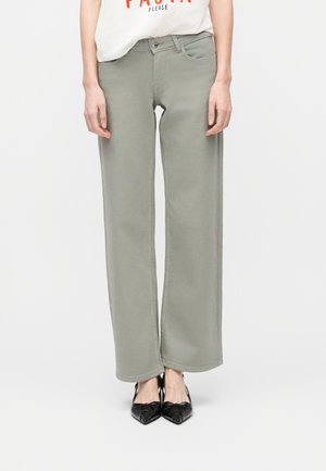 Žena oblečená v svetlosivých širokých nohaviciach, čierne špicaté balerínky a bielu skrátenú blúzku s oranžovým textom, stojaca na bielom pozadí.