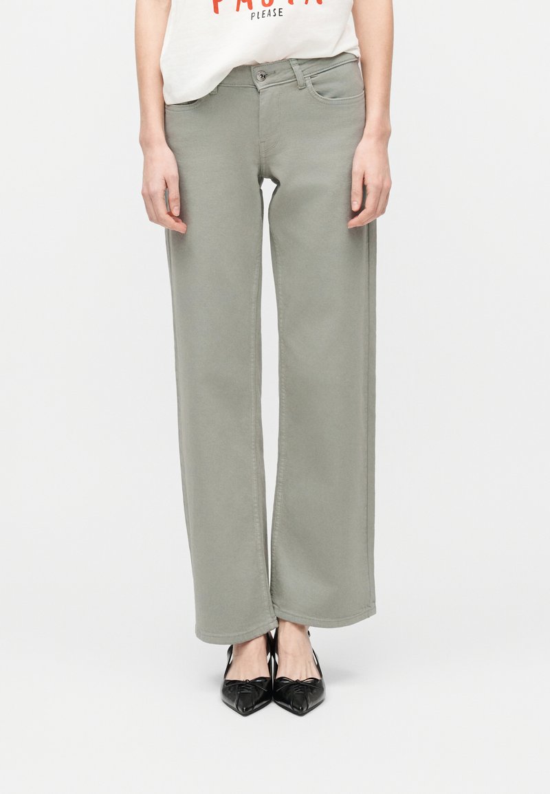 Γυναίκα που φοράει ανοιχτογκρι παντελόνια με φαρδιά πόδια, μαύρα φλατ παπούτσια με μυτερή μύτη και ένα λευκό κοντό πουκάμισο με πορτοκαλί κείμενο, στέκεται μπροστά σε λευκό φόντο.