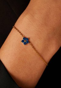 Gouden armbandschakel met een blauw bloemenontwerp, voorzien van vijf ronde stenen en een centrale gouden accent. Gladde textuur tegen de huid.