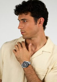 Orologio da polso argentato con quadrante nero, numeri marcati e cinturino testurizzato. Il modello indossa una camicia color crema con colletto rilassato.