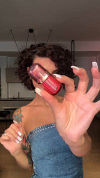 Rote Glas-Kosmetikflasche mit metallic-goldenem Akzent, gehalten von einer Hand mit langen N�ägeln, vor einem neutralen Indoor-Hintergrund.