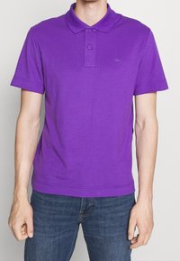 Camisa polo morada de algodón, con cuello clásico, tapeta de dos botones y mangas cortas. Combinada con vaqueros azul oscuro.