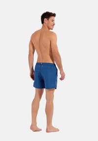 Shorts de bain bleus à motif en losanges, avec poches latérales et une taille à bouton. Le modèle est pieds nus et vu de dos.
