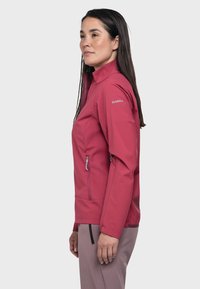 Rote Softshelljacke mit langen Ärmeln, hohem Kragen und zwei Reißverschlusstaschen. Mit einem taillierten Design und einem Logo auf der Schulter.