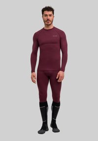 Bordowy termiczny zestaw bielizny podstawowej, składający się z dopasowanej bluzki z długim rękawem i dopasowanych spodni. Noszony z czarno-szarymi skarpetami sięgającymi do kolan.
