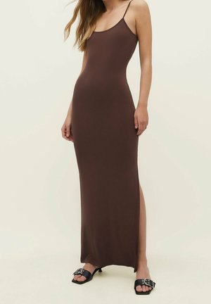 Vrouw in een mouwloze, bruine maxi-jurk met een zijsplit en zwarte open-toe sandalen met gespdetails, staande tegen een effen achtergrond.