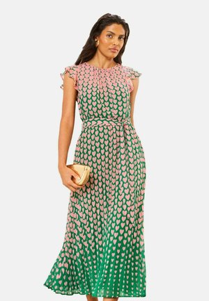 Kvinde i grøn midi-kjole med pink hjerteprint, flagrende ærmer, bindebånd i taljen, holder en guldbelagt clutch.