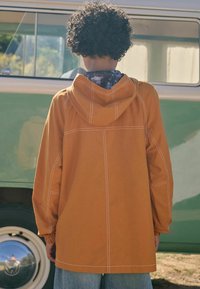 Orange huva jacka med vit sömnad, lös passform och långa ärmar, parat med ljusblå denimjeans. Bakvyn visar texturerad tyg.