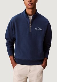 Sudadera azul marino con cremallera, cuello alto y logo "calvin" bordado. Fabricada con tejido suave, presenta un corte relajado y puños acanalados.