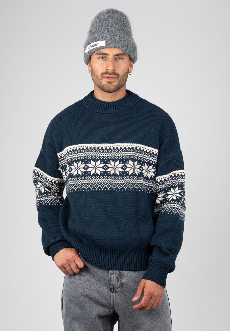 Homme portant un bonnet en laine gris et un pull bleu marine avec des motifs de flocons de neige blancs, debout devant un fond clair uni.