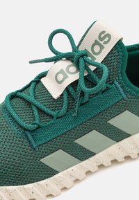 Grön nät-sneaker med texturerad överdel, snörning, stor logotab, och gummisula med ett fläckigt mönster. Design med tre ränder.