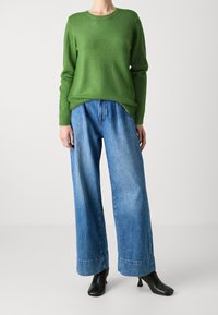 Grüner Wollpullover mit rundem Ausschnitt, kombiniert mit locker sitzenden blauen Jeans aus Denim mit breiten Säumen. Schwarze, hochhackige Stiefeletten runden das Outfit ab.