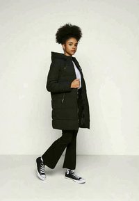 Abrigo acolchado negro con capucha y cremallera frontal, con cintura ajustada y costuras horizontales. Se lleva con una camisa blanca y pantalones negros, combinados con zapatillas negras.