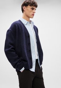 Tengerész kék kardigán, textúrázott kötésű, fekete gombokkal és bordás mandzsettával, világoskék csíkozott inggel viselve; laza pózban bemutatva.