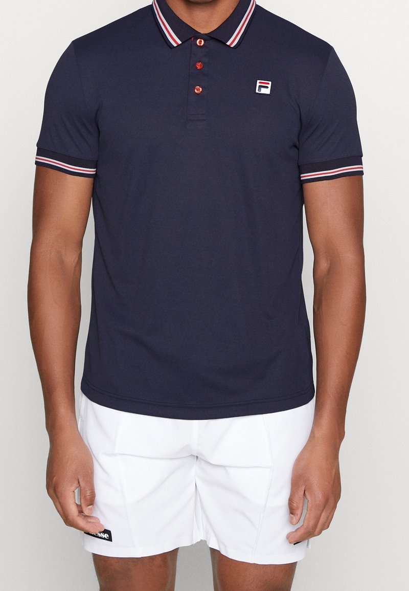 Polo bleu marine en tissu lisse, avec un col et des bordures de manches rayés en rouge et blanc, arborant un logo sur la poitrine.