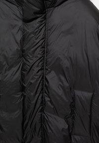 Czarna, pikowana tkanina z pionowym szwem i małą naszywką, ukazująca strukturę oraz delikatne refleksy światła.