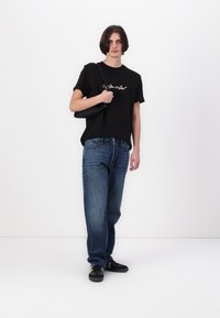 Camiseta de algodón negra con logo blanco, combinada con jeans de mezclilla azules. El modelo lleva un bolso cruzado negro y usa zapatillas negras.