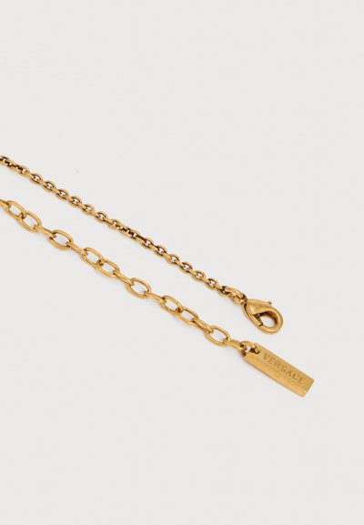 Αλυσίδες από μέταλλο σε χρυσαφί απόχρωση με κούμπωμα τύπου καβούρι, μία πιο χοντρή αλυσίδα με ορθογώνια ταμπέλα χαραγμένη με την ένδειξη «VERSACE» σε λευκό φόντο.