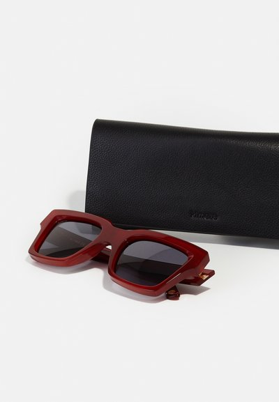 Ochelari de soare supradimensionați roșii, cu lentile întunecate, având un design geometric al ramei. Însoțiți de o carcasă din piele neagră texturată.