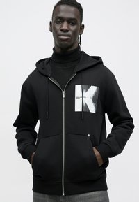 Sweatshirt preto com capuz e fecho de correr frontal, com um logótipo branco com linhas verticais. Possui um bolso canguru e punhos canelados.