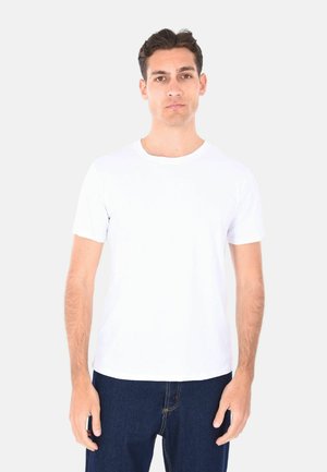 Giovane uomo con capelli scuri che indossa una semplice t-shirt bianca a maniche corte e jeans blu scuro, in piedi davanti a uno sfondo grigio chiaro.