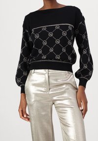 Pull noir en tricot avec texture côtelée, présentant un motif géométrique de logo argenté et des manches bouffantes. Associé à un pantalon argenté métallisé brillant.