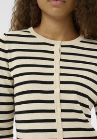 Ženska, oblečena v bež kardigan s gumbi in črnimi vodoravnimi črtami, z kodrastimi lasmi, ki so vidni do ramen.