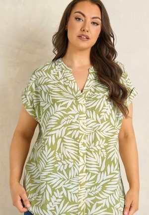 Kvinde med langt brunt hår iført en grøn bluse med korte ærmer og hvide bladmønstre, stående mod en neutral baggrund.