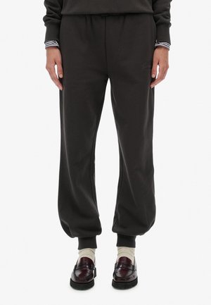 Parte inferior de una persona que lleva pantalones jogger negros, puños de camisa a rayas, calcetines blancos y mocasines de cuero rojo oscuro sobre fondo blanco.