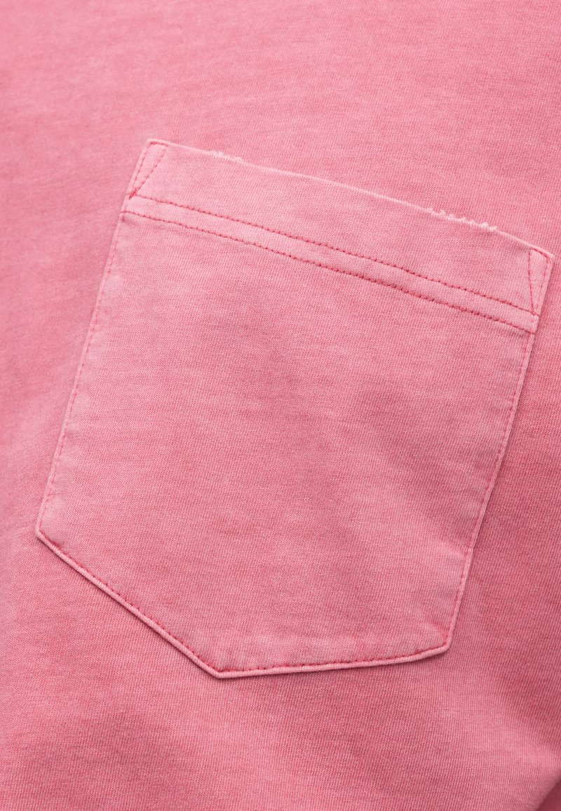 Lyserødt bomuldsstof med en påsyet lomme. Blød tekstur, let falmet udseende, med dobbeltsyning langs lommens kant.