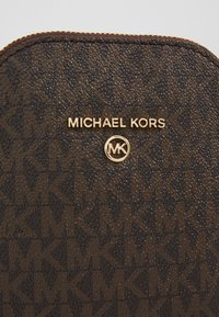 Bolso de mano marrón con textura que presenta un patrón geométrico, con el logotipo "Michael Kors" en tono dorado y un emblema circular "MK" en la parte frontal.