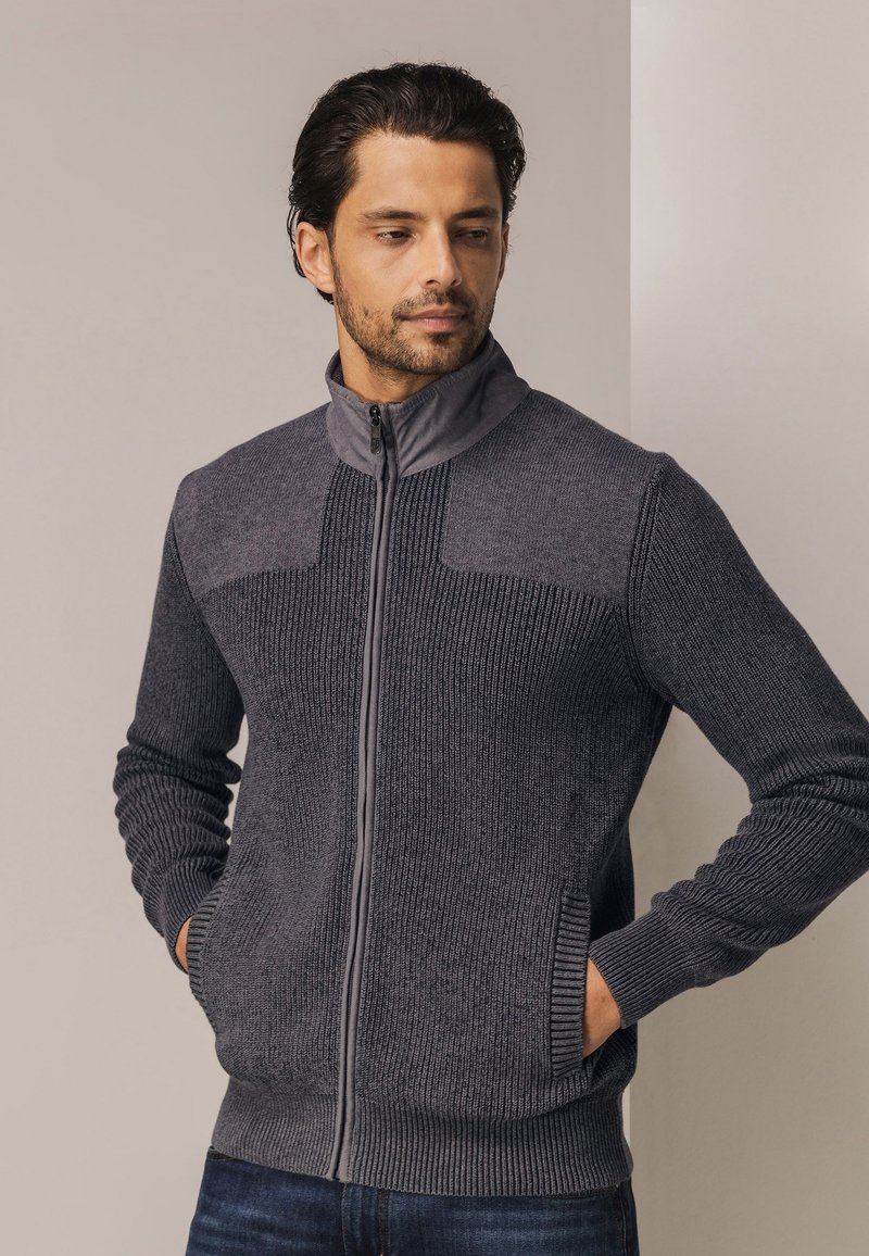 Donkergrijze ritstrui met geribbelde textuur, contrasterende lichtgrijze schouders en zijzakken. Heeft een hoge kraag en aansluitende manchetten.