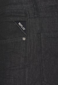 Jeans de mezclilla negra con un diseño ajustado, que presentan costuras blancas, un botón de metal y una etiqueta tejida que muestra "REPLAY" en el bolsillo.