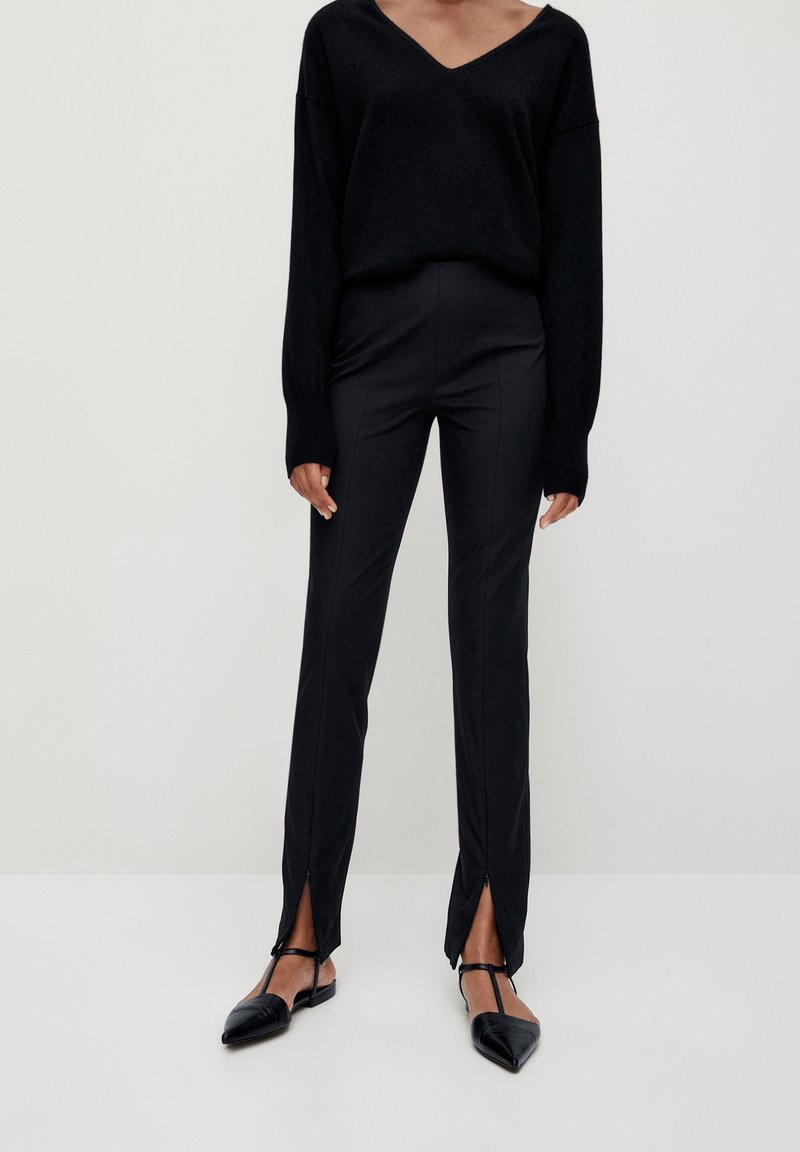 Femme portant un pull noir à col en V, un pantalon noir ajusté avec des fentes à l'avant aux chevilles, et des ballerines noires à bride en T à bout pointu, debout sur un sol blanc.