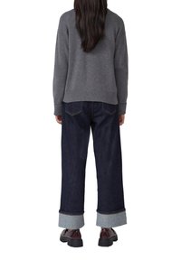 Vrouw die weggedraaid staat met donkere opgerolde jeans, een grijze trui en donkerbruine schoenen met een dikke zool tegen een witte achtergrond.