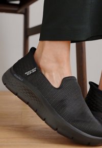 Black slip-on shoes with a textured mesh upper, elasticated collar, and cushioned sole. Simple design with Skechers branding on the side.