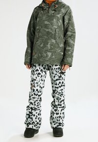 Grön kamouflagejacka med huva och två framfickor, kombinerad med byxor i vit och svart leopardmönster. Svarta snöboots fulländar outfiten.