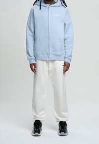 Hoodie zippé bleu clair avec poches avant et logo blanc, associé à un pantalon blanc ample. Porté avec des chaussures de sport noires et grises.