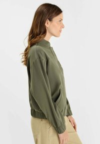 Femme aux longs cheveux bruns portant une veste à manches longues vert olive avec des poignets élastiques et un pantalon beige, de profil face à droite.