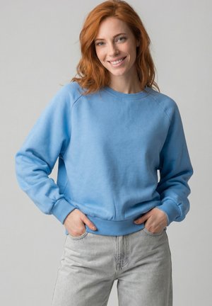 Junge Frau mit roten Haaren lächelt, trägt ein hellblaues Sweatshirt und hellgraue Jeans, steht mit den Händen in den Taschen vor einem schlichten Hintergrund.