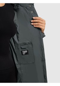 Vêtements d'extérieur matelassés verts avec fermeture à pression et poche latérale. L'étiquette affichée indique "Khujo original for a few", mettant en valeur un tissu durable et une texture.