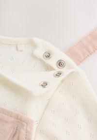 Close-up of a cream knitted garment with small holes and metal snap buttons near a pink ribbed fabric strap.