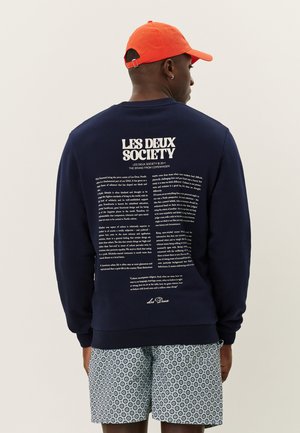 Sudadera de cuello redondo azul marino con diseño de texto en la parte posterior. Puños y dobladillo acanalados. Combinada con pantalones cortos con estampado y una gorra naranja.